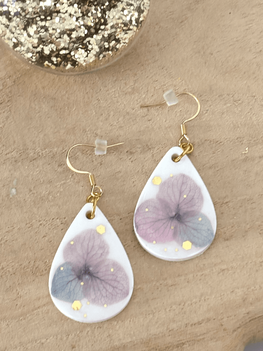 Paire de boucles d'oreilles en goutte blanche avec fleur d'hortensia mauve posées sur du bois.
